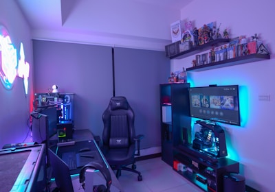 A modern gaming setup featuring a sleek gaming chair, dual monitors on a desk, and LED lighting. A gaming console and various decorations are displayed on shelves, creating an organized and vibrant gaming environment.