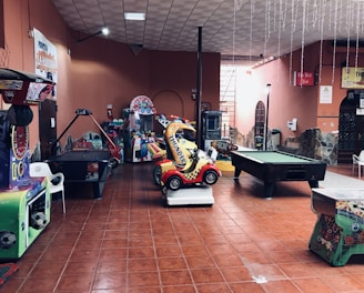 An indoor arcade area featuring various gaming machines, including an air hockey table and a ride-on toy car. The room has a tiled floor and several colorful gaming devices scattered around. Chairs and arcade signage are visible, creating a playful and entertainment-focused atmosphere.